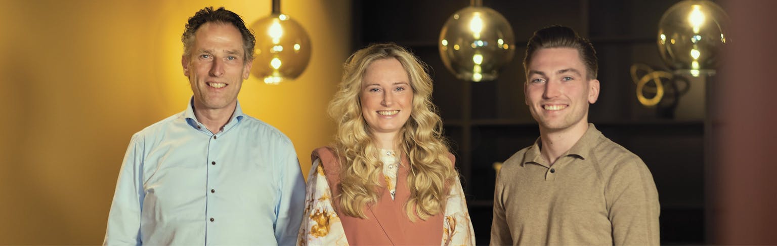 Drie mensen poseren op een foto. Ze staan voor een gele wand en ronde lampen. Links staat een man met en lichtblauw overhemd, in het midden een vrouw met blond haar en een bruinroze pak aan. Rechts staat een man met een zandkleurige trui. 
