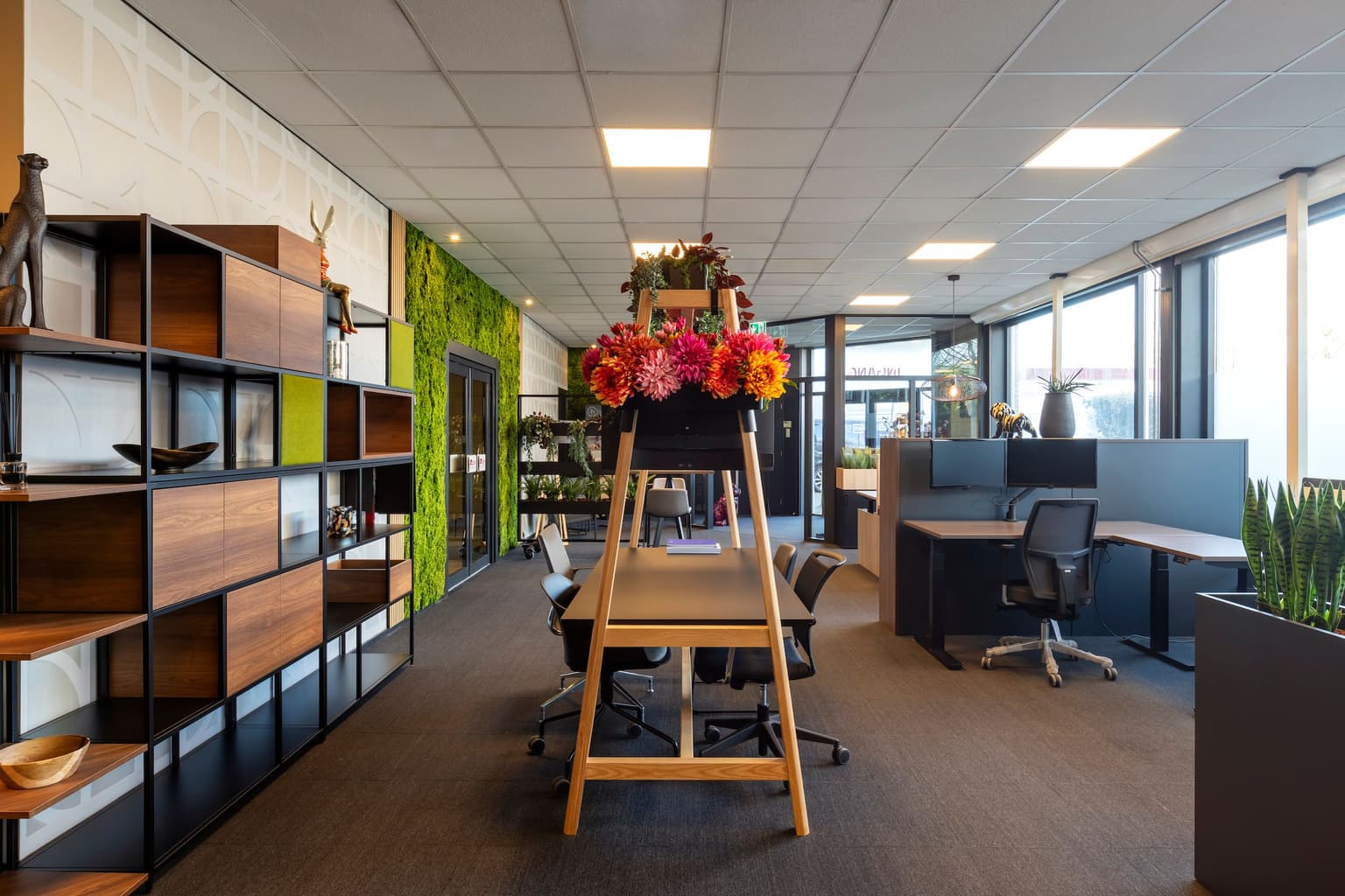 Kantoorruimte met links tegen de wand, een open wandkast. Verderop een groene moswand. In het midden een vergadertael met ombouw. Daaraan hangen kunstplanten en zijden bloemen. Rechts zien we op hoogte verstelbare werkplekken. 