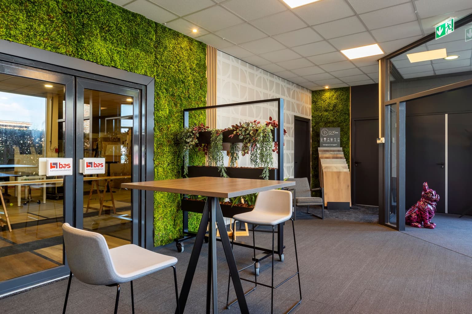 Kantoorruimte van BAS. Links zien we de moswand, met een deur naar de showroom. Er staat een verrijdbare plantenbak, gevuld met kunstplanten. En in de voorgrond staat een bartafel met twee barstoelen eromheen. 