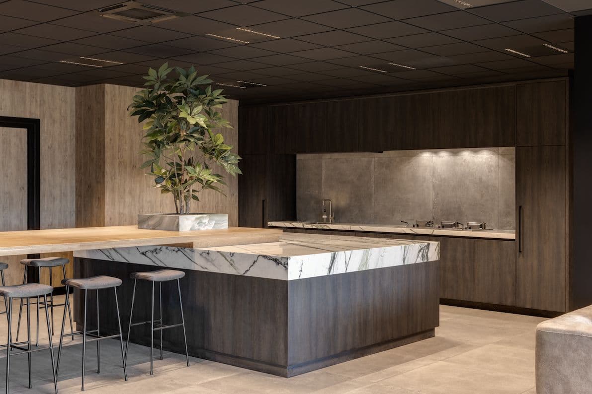 Kookeiland in de kantine bij Ansova. Deze is van donker hout met een marmeren blad erop. Aan het eiland zit een bar vast met barstoelen. 