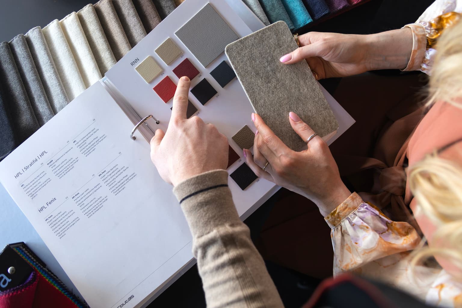 Een mannenhand wijst een tegeltje aan in een stalenboek. Twee vrouwenhanden houden een petvilten staal vast in de kleur grijs. 