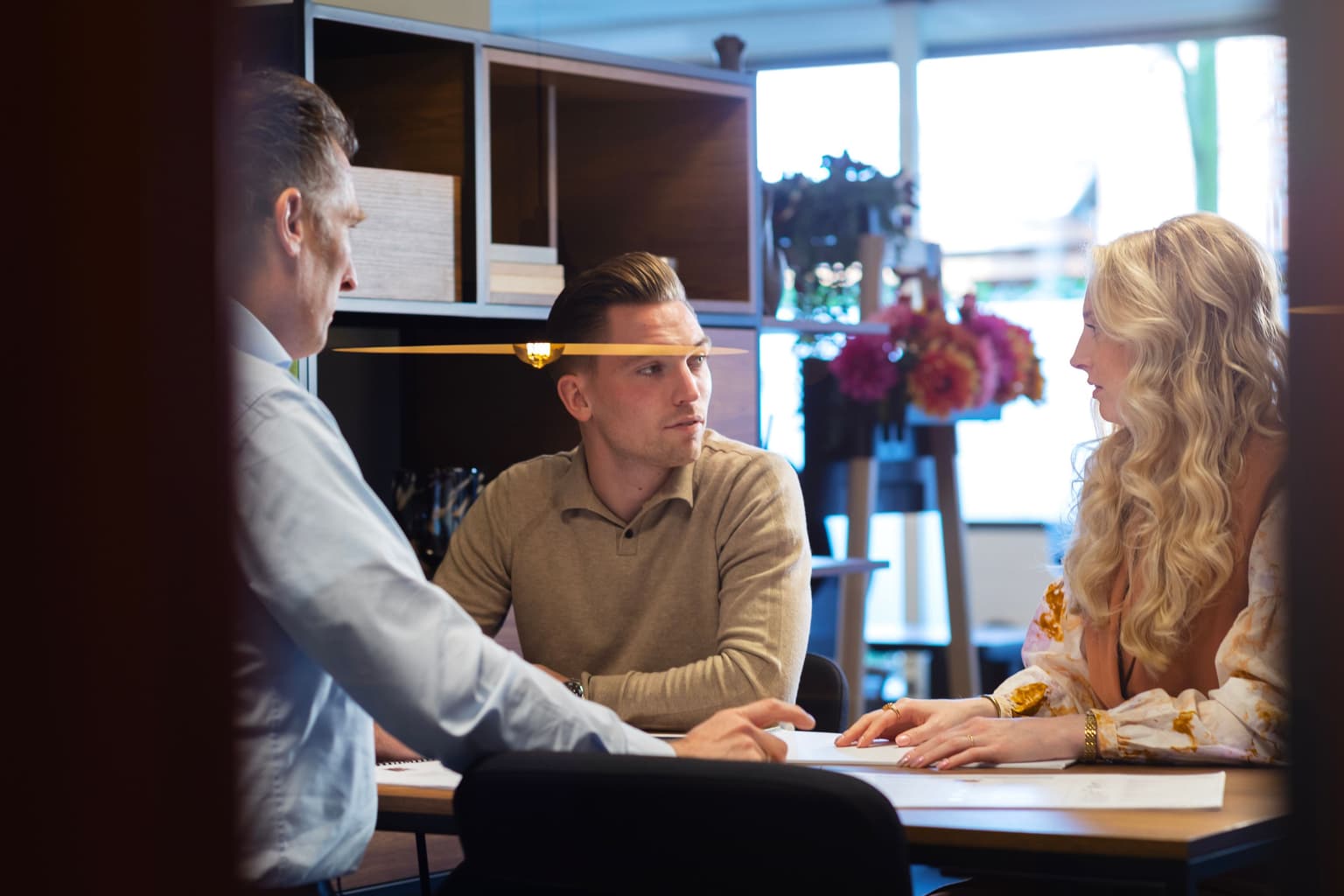 Joan kijkt naar Anne en zit links aan de tafel. Anne zit rechts van hem. Casper staat met de rug naar ons toe. In de weerspiegeling van het glas zien we een lamp schijnen. 