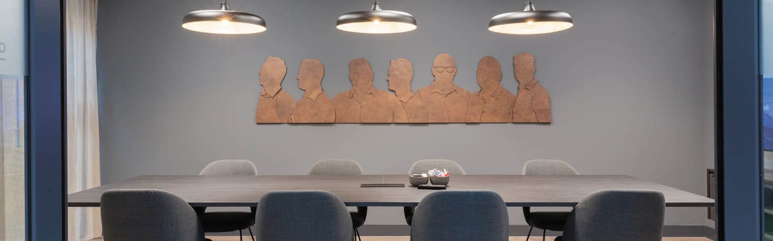Vergaderruimte met grijze muren, vergadertafel met donkerblad en acht grijze vergaderstoelen er langs. Er hangen drie smalle lampen boven de tafel en op de muur is een bronzen uitsnede van de silhouetten van de familie. 