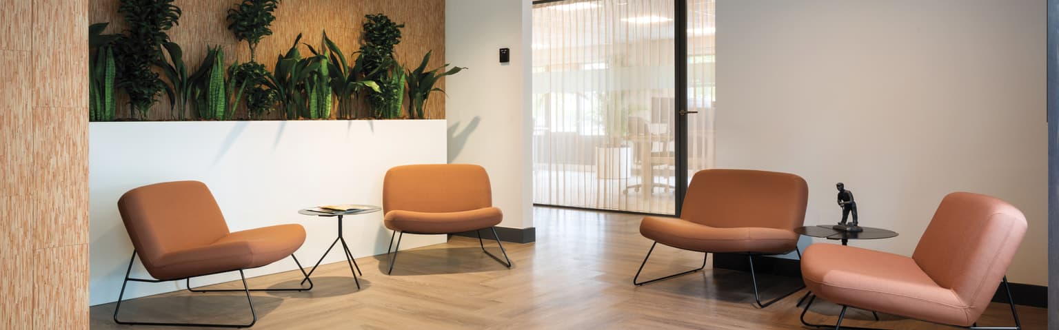 Wachtruimte met daarin een lichte visgraatvloer, bruine leren wachtkamerstoelen. Links zien we een witte plantenbak met hoge groene planten erin. 