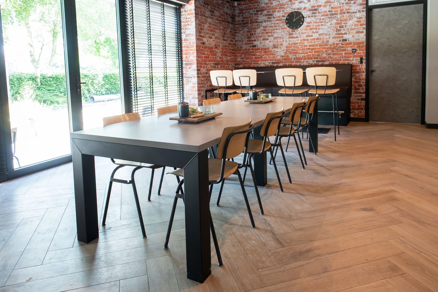 Kantine met een visgraatvloer, eettafel met ijzerenframe en betonlook blad, met simpele houten kantine stoelen en tegen de muur aan een bartafel met houten barkrukken. De deuren hebben betonlook.