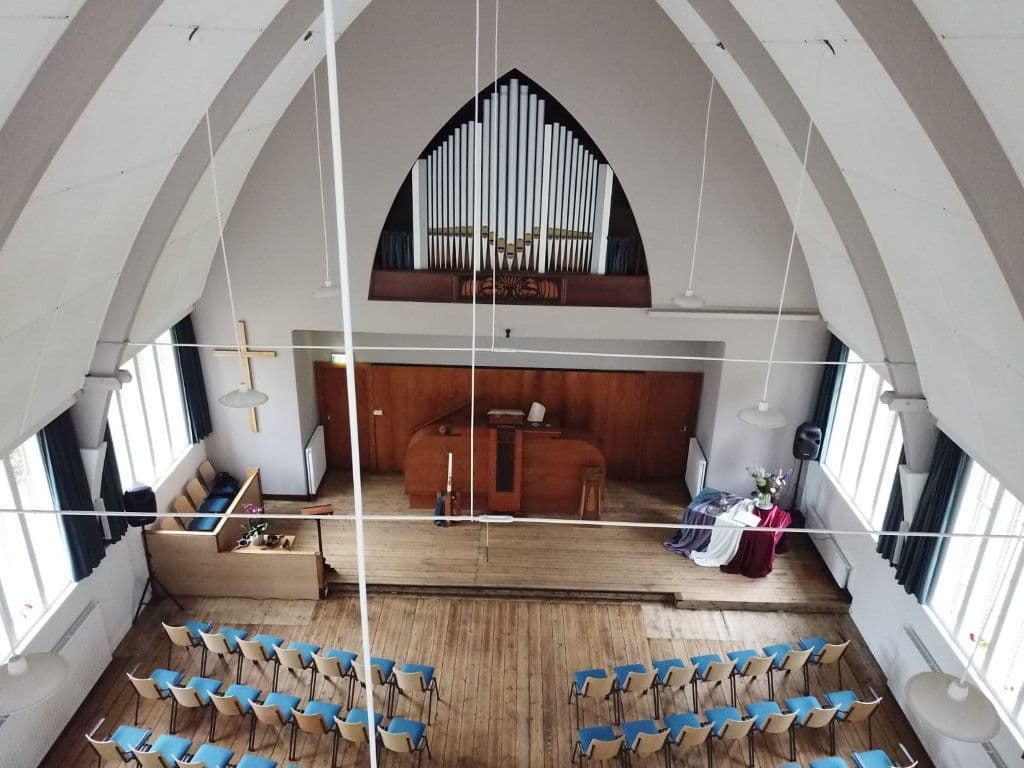 Van bovenaf kijken we op de kerk neer. We zien de rijen met kerkstoelen met lichtblauwe zittingen, de kansel, een gouden kruis tegen de muur en een driehoekig kerkorgel.