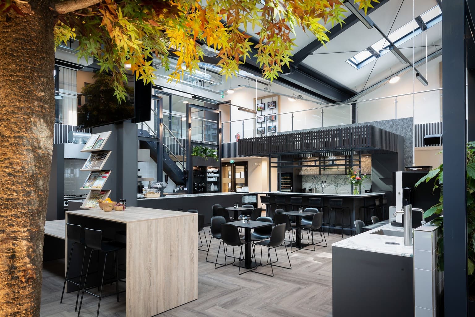 Kantine bij Den Ouden Groep, met in de voorgrond een boom, een overkapte bar en in het midden zwarte vierkante tafeltjes, met daaromheen zwarte stoelen.