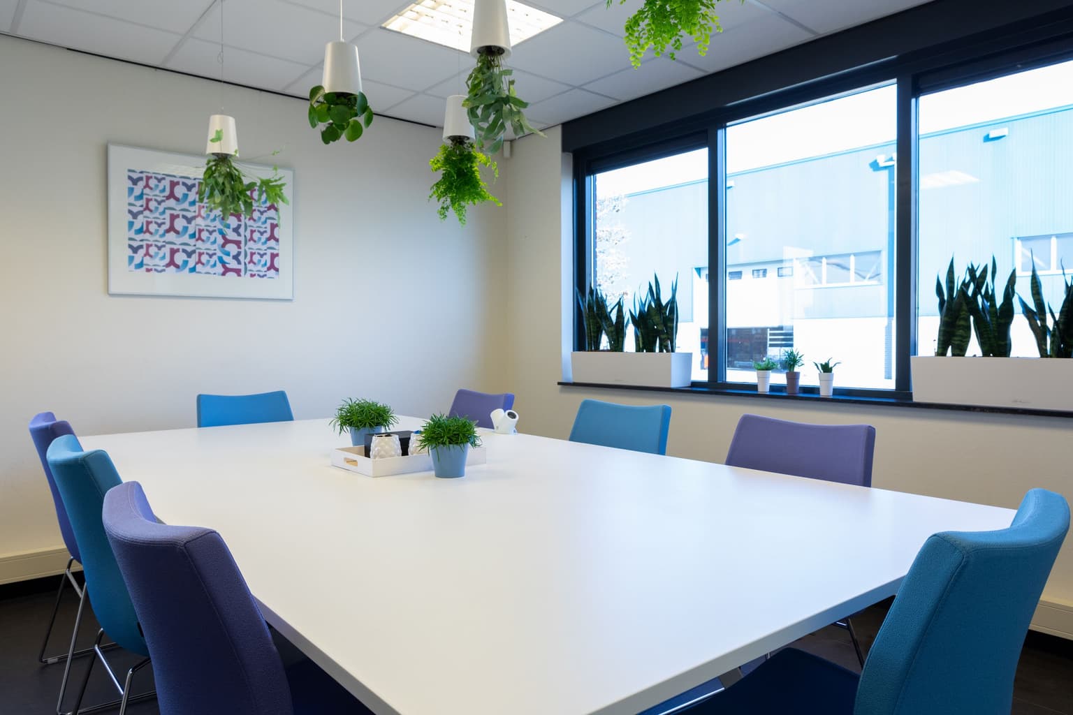 Vergaderruimte met een witte vergadertafel en daaromheen paarse en turqoise vergaderstoelen. Boven de tafel hangen planten.
