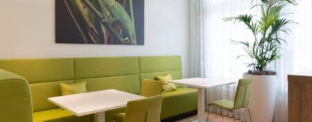 Kantine met daarin groene treinbanken, twee witte tafels met twee groene stoelen aangeschoven en rechts in de hoek een witte pot met een grote plant.