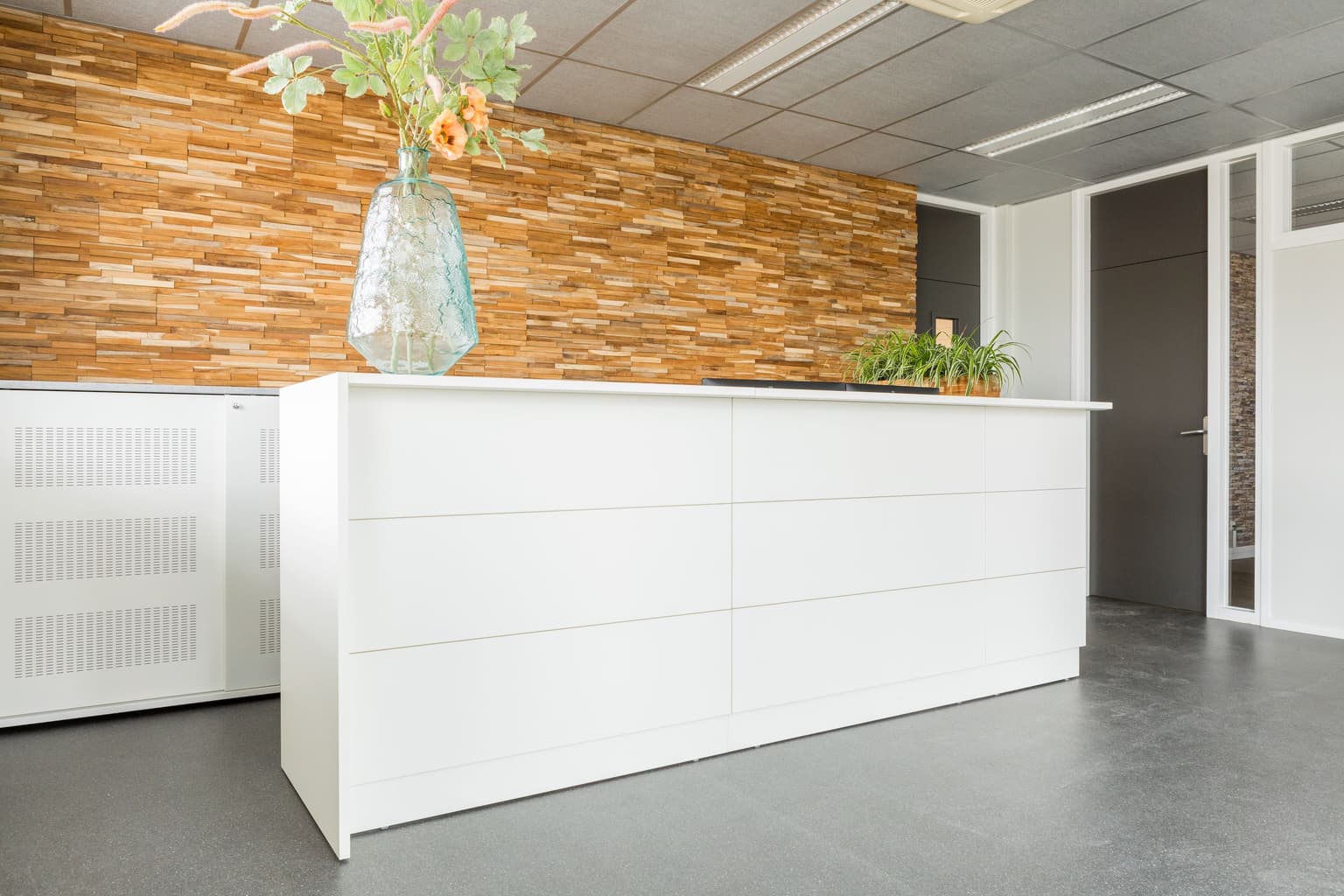 Witte receptiebalie met daarachter een houten wand en op de balie staat een grote vaas met kunstbloemen erin.