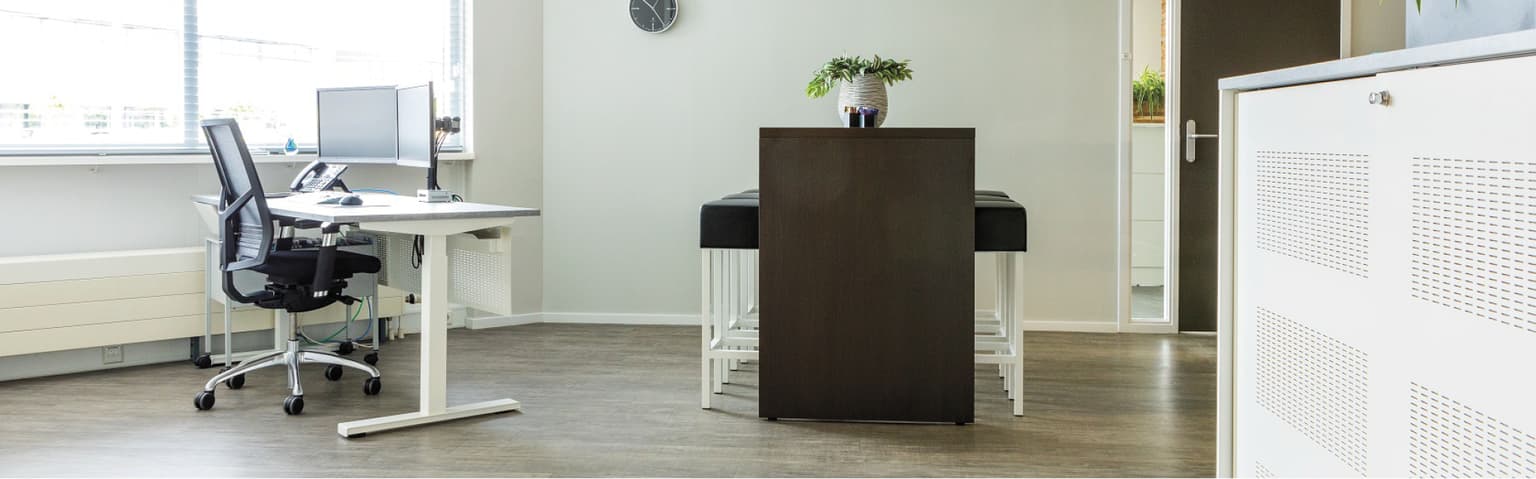 Links een werkplek met een wit zit-stabureau en een zwarte bureaustoel en een dubbele monitorarm. Rechts staat een bartafel met zwarte barkrukken eraan.