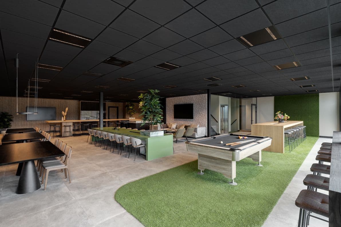 Kantine bij Ansova Staalcoating. Daarin staat een lange kantinetafel, vierkante kantinetafels, een hoge kantinetafel en een pooltafel. Daaronder ligt een grasmat.