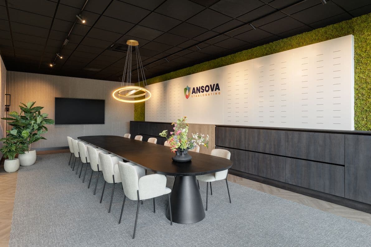 Vergaderruimte met een vergadertafe voor 12 personen erin. Rechts aan de muur zien we een moswand met het logo erop. Boven de tafel hangt een designlamp met ringen.