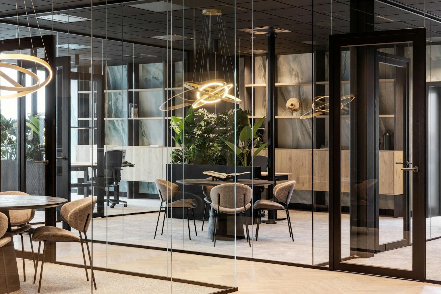 Vergaderruimte met een glazenwand, een ronde vergadertafel en zandkleurige stoeltjes met een designlamp erboven.