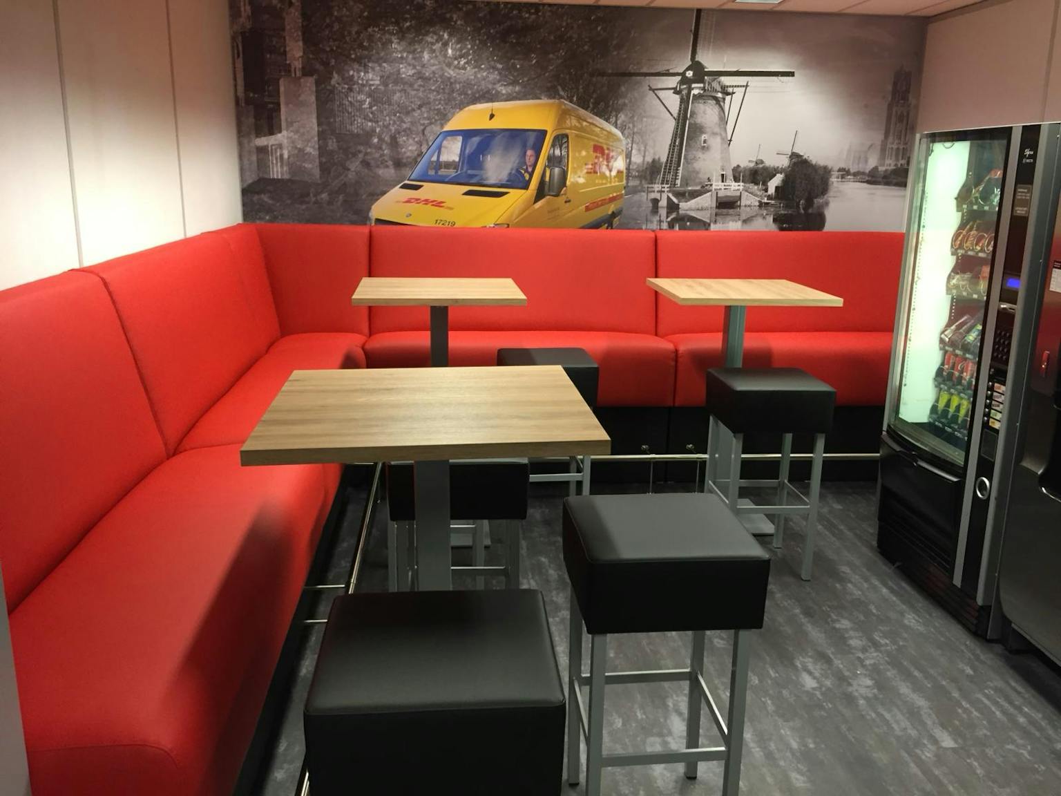 Koffiecorner met rode treinbanken die de hoek vullen. Voor de treinbanken staan bartafels met lichteiken houten bladen en daarbij staan weer zwarte krukken. Op de muur is een visual geplakt met een DHL-bus.