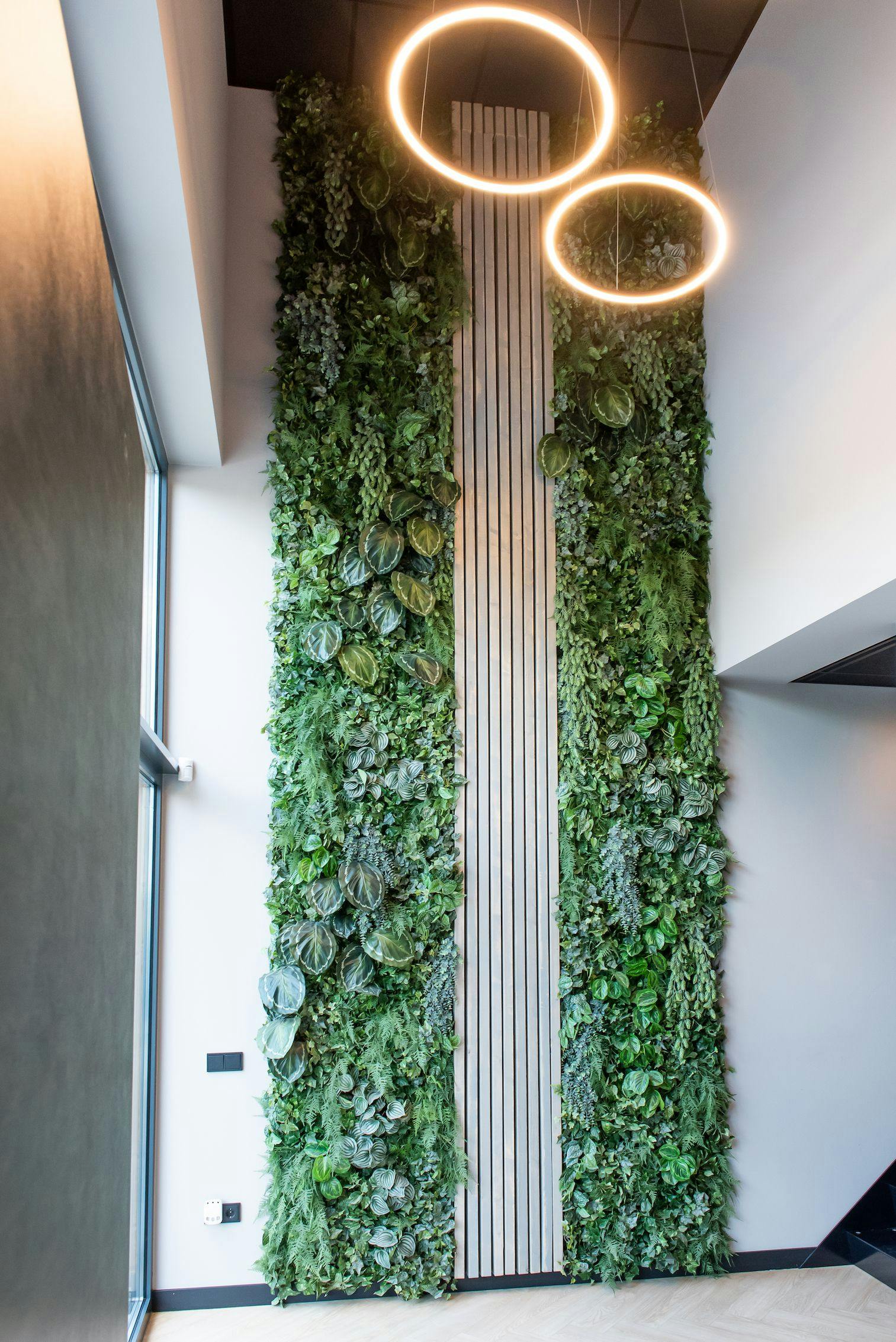 Entree van een kantoorpand, de hoge wand is bekleed met planten en in het midden met houten latten. Aan het plafond hangen twee cirkellampen.