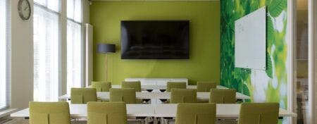 Vergaderruimte met links een glazen pui en voorin een groene muur, met daar een tv tegenaan. Parallel aan de groene muur staan witte tafels met groene stoelen.