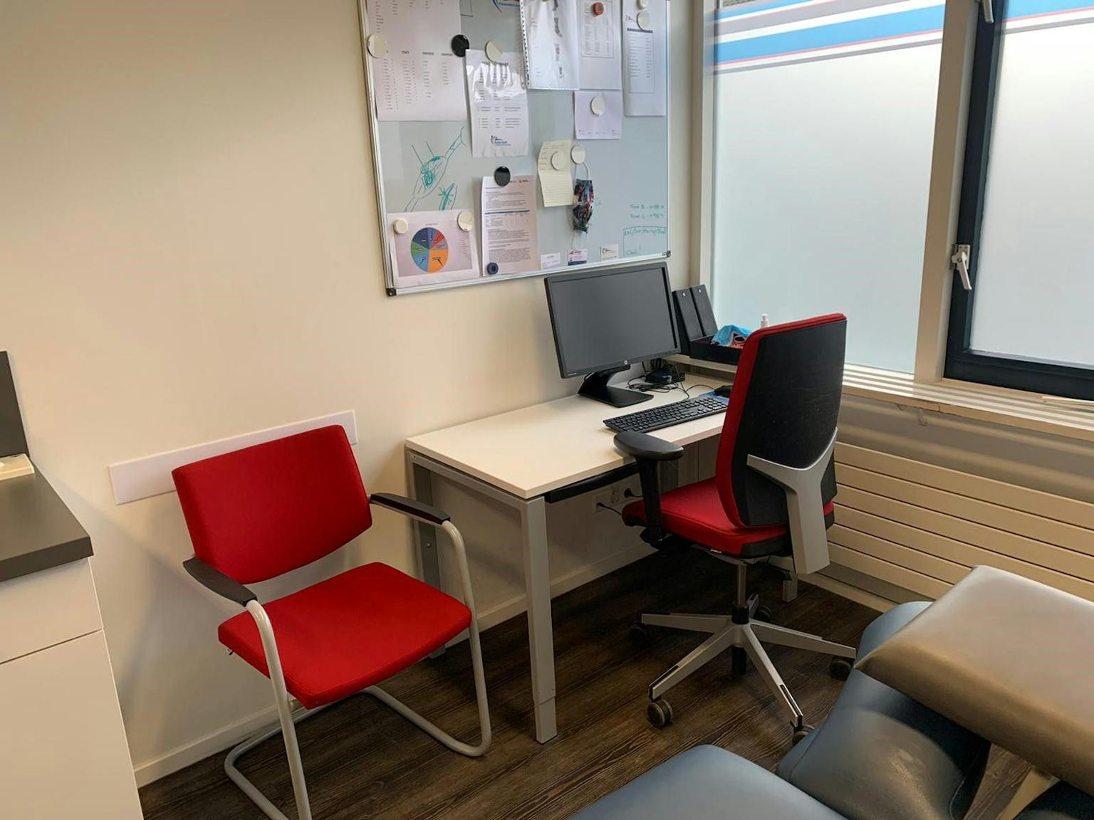 Spreekkamer met daarin een wit bureau, daarachter een rode bureaustoel en naast deze werkplek staat een rode stoel voor patiënten.