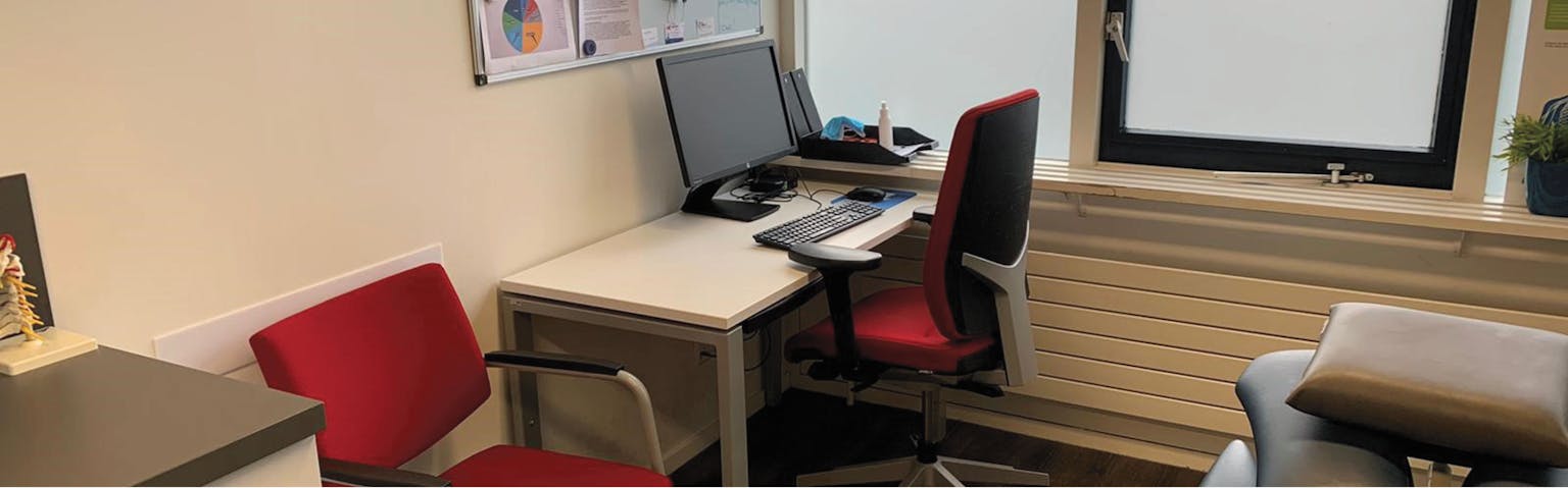 Spreekkamer met daarin een wit bureau, daarachter een rode bureaustoel en naast deze werkplek staat een rode stoel voor patiënten.