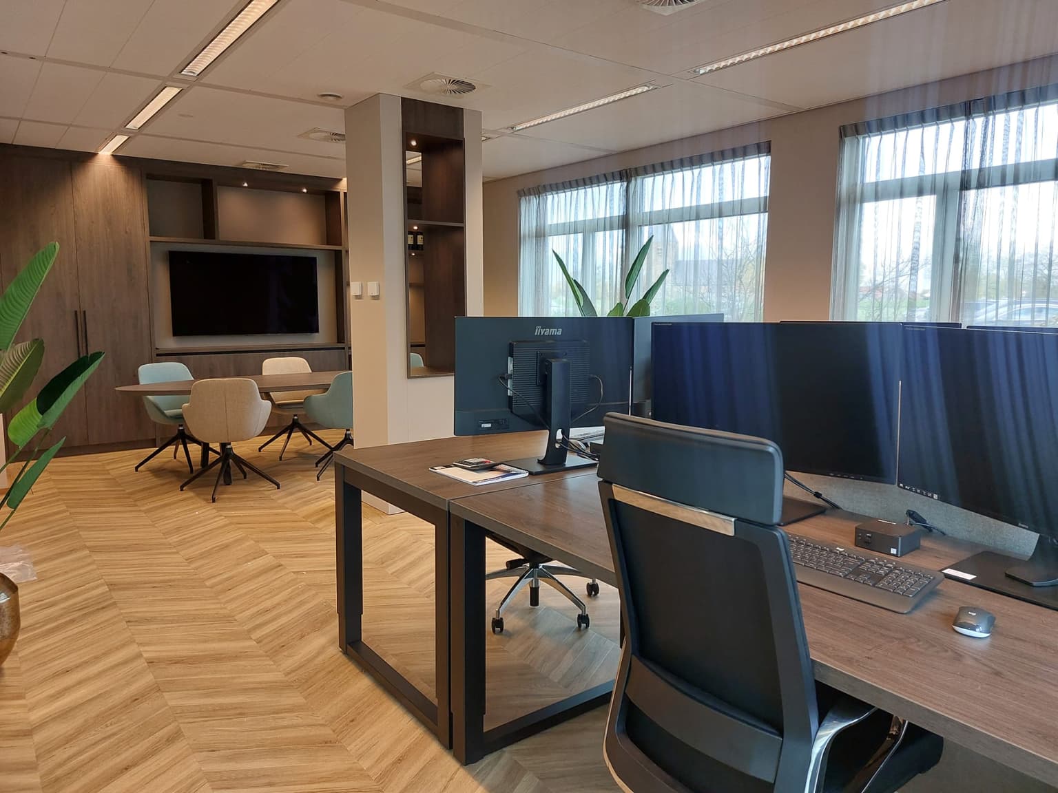 Kantoorruimte met een hongaarse punt-vloer. Achterin staat een vergadertafel met een groot beeldscherm erachter. In de voorgrond staan werkplekken van donker hout.